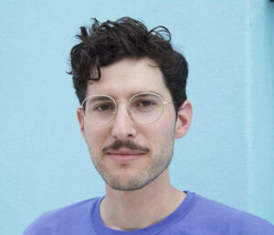 Headshot of a man wearing a violet t shirt and a spectacle