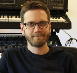 Headshot of Adam Fligsten in black t shirt wearing specs